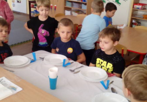 Czterej chłopcy siedzący przy nakrytym przez nich stole. Dwóch stojących chłopców ocenia poprawność nakrycia.